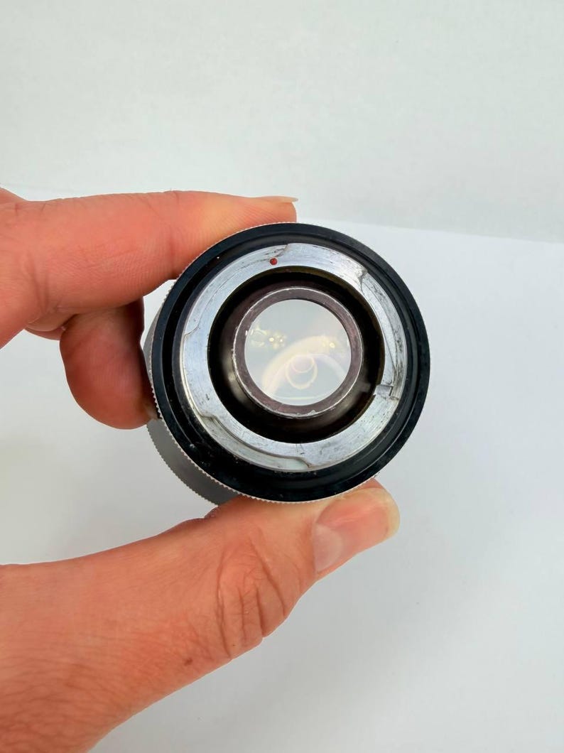 Puede incluir: Primer plano de una lente de c&aacute;mara, con un anillo de metal plateado y un agarre de goma negro. La lente tiene un elemento de vidrio transparente en el centro. Un peque&ntilde;o punto rojo es visible en el anillo de metal.