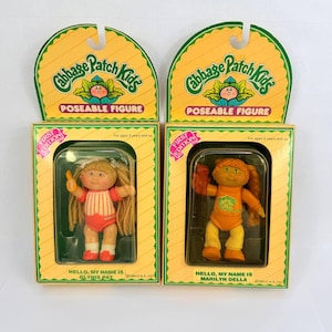 Op de afbeelding: Twee vintage Cabbage Patch Kids poseerbare figuren in hun originele dozen. De dozen zijn geel met groene randen en tonen het Cabbage Patch Kids logo. Eén pop is Glynis Pat, de andere is Marilyn Della. Elke pop draagt een kleurrijke outfit.