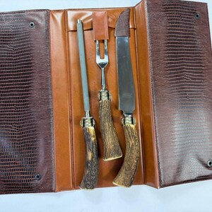 May include: A brown leather case with a reptile skin texture, open to reveal a sharpening steel, carving fork, and carving knife. The handles are made of a brown antler material with brass accents. The case is brown and tan.