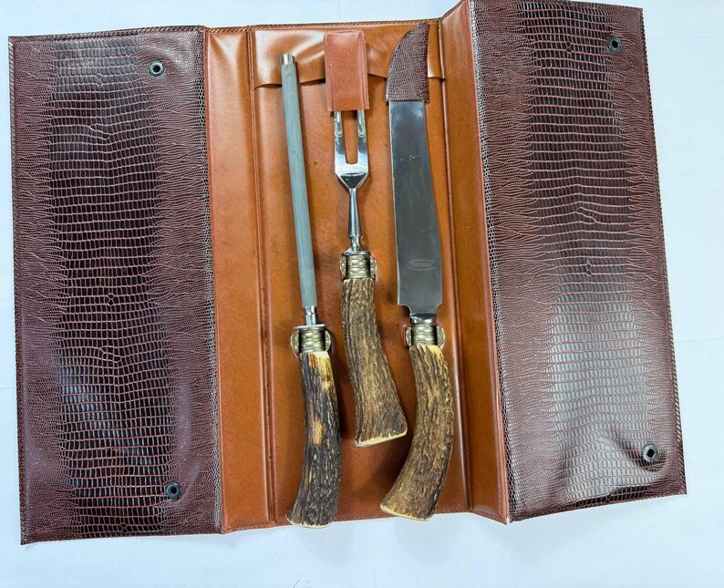 May include: A brown leather case with a reptile skin texture, open to reveal a sharpening steel, carving fork, and carving knife. The handles are made of a brown antler material with brass accents. The case is brown and tan.