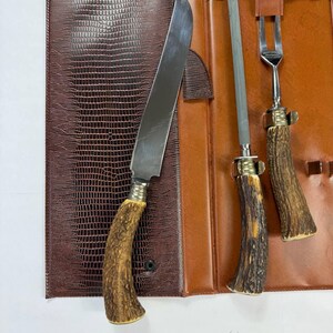 May include: A brown leather knife set with a knife, fork, and sharpening steel. The knife and fork have antler handles. The set is in a brown leather case with a textured, reptile-like pattern.
