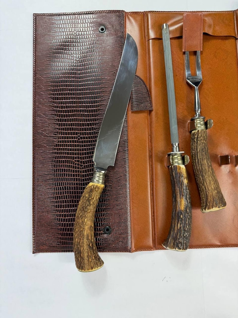 May include: A brown leather knife set with a knife, fork, and sharpening steel. The knife and fork have antler handles. The set is in a brown leather case with a textured, reptile-like pattern.