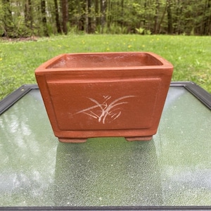 Op de afbeelding: Een vierkante bonsai pot in terracotta kleur met een wit, gestileerd bloemmotief. De pot heeft een licht taps toelopende vorm en staat op vier kleine voetjes. De achtergrond toont een wazig zicht op een groen gazon en bomen.