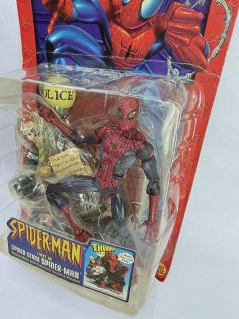 Peut inclure: Figurine articul&eacute;e de Spider-Man dans un emballage plastique. La figurine est rouge et noire, avec un costume d&eacute;taill&eacute;. L'emballage pr&eacute;sente le logo Spider-Man et le texte "Spider-Sense Spider-Man". L'arri&egrave;re-plan montre une illustration de Spider-Man.