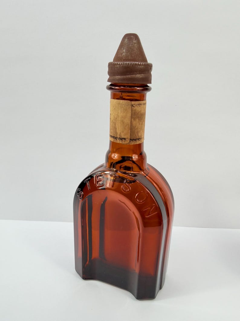 Puede incluir: Una botella de vidrio &aacute;mbar con una tapa de metal marr&oacute;n. La botella tiene forma rectangular con hombros redondeados y la palabra "STETSON" en relieve en la parte delantera. Una etiqueta de papel descolorida est&aacute; adherida al cuello.