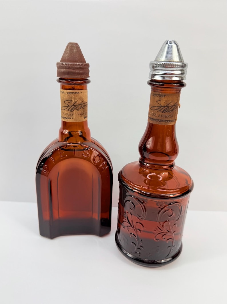 Puede incluir: Dos botellas vintage de vidrio &aacute;mbar. Una es rectangular con tapa marr&oacute;n, la otra cil&iacute;ndrica con tapa plateada y dise&ntilde;o floral en relieve. Ambas tienen etiquetas que dicen "Stetson".