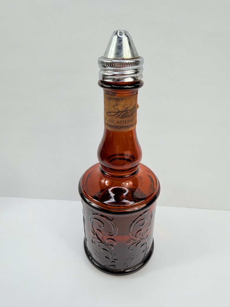 Puede incluir: Una botella de vidrio marr&oacute;n vintage con una tapa de metal plateado. La botella tiene un dise&ntilde;o en relieve decorativo y una etiqueta que dice "Ste... 2 OZ AFTER". La botella es probablemente para aftershave o colonia.