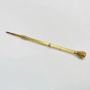 Könnte beinhalten: Ein goldfarbener Druckbleistift mit spitzer Spitze und Zierdetails. Der Bleistift hat einen strukturierten Schaft und eine dekorative Endkappe. Der Bleistift liegt auf einer weißen Oberfläche.
