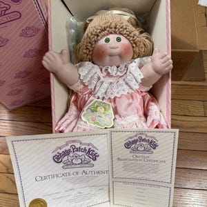 Può includere: Una bambola in porcellana Cabbage Patch Kids con capelli di lana, occhi verdi e guance rosate, che indossa un abito di raso rosa con bordo in pizzo bianco. La bambola è in una scatola rosa con un certificato di autenticità e di registrazione.