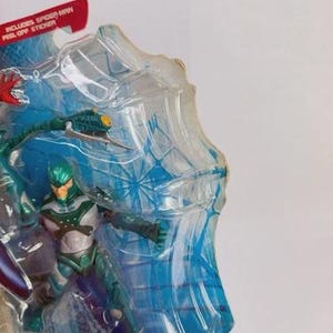 Puede incluir: Una figura de acci&oacute;n empaquetada de un personaje con un traje verde azulado y plateado con un casco verde. El embalaje incluye el texto "Includes Spider-Man Peel-Off Sticker" y "Tail Shoots Water!"