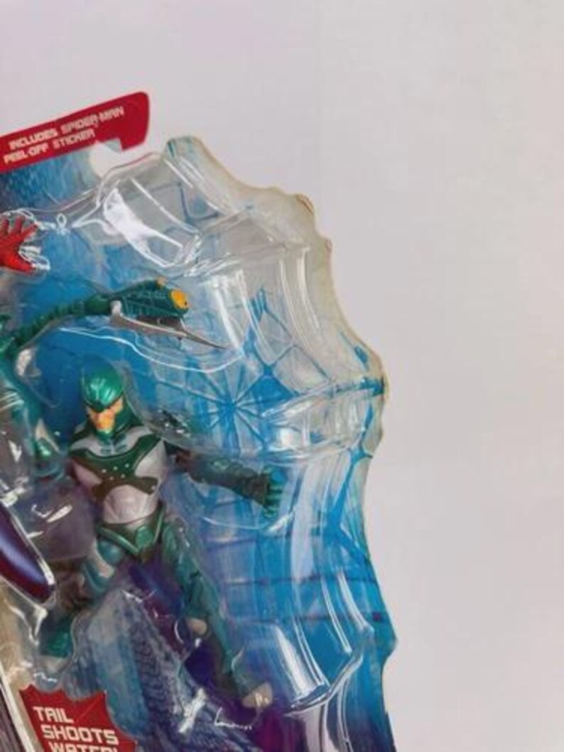 Puede incluir: Una figura de acci&oacute;n empaquetada de un personaje con un traje verde azulado y plateado con un casco verde. El embalaje incluye el texto "Includes Spider-Man Peel-Off Sticker" y "Tail Shoots Water!"