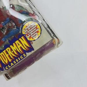 May include: A Spider-Man action figure in a clear plastic package. The figure is red and blue, with the words "SPIDER-MAN CLASSICS" in a yellow and blue logo. The package includes a collector's book and a wall-mountable display stand.