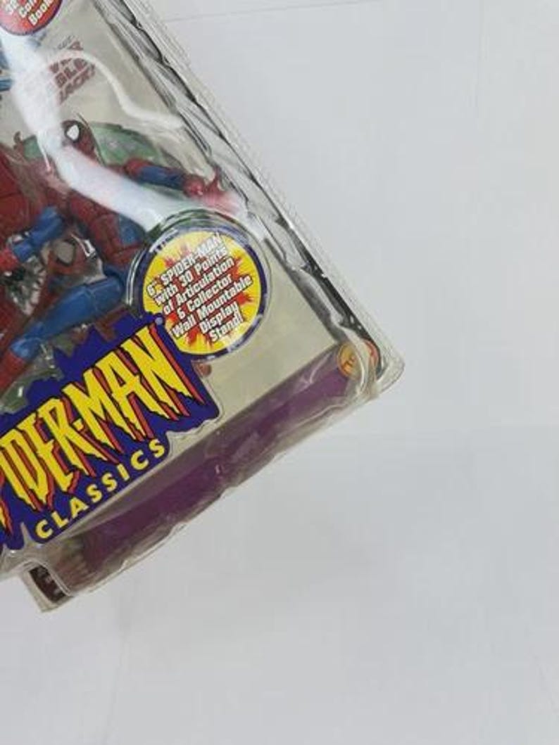 May include: A Spider-Man action figure in a clear plastic package. The figure is red and blue, with the words "SPIDER-MAN CLASSICS" in a yellow and blue logo. The package includes a collector's book and a wall-mountable display stand.