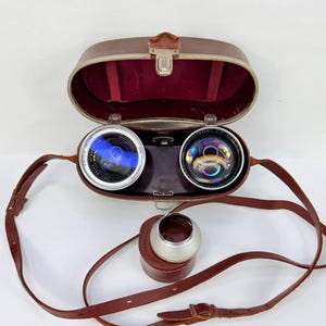 Puede incluir: Lentes de c&aacute;mara vintage con estuche y correa de cuero. Las lentes son plateadas y negras, con un interior azul reflectante. El estuche es marr&oacute;n con interior de terciopelo rojo. Tambi&eacute;n se incluye un parasol plateado y un peque&ntilde;o estuche de cuero.