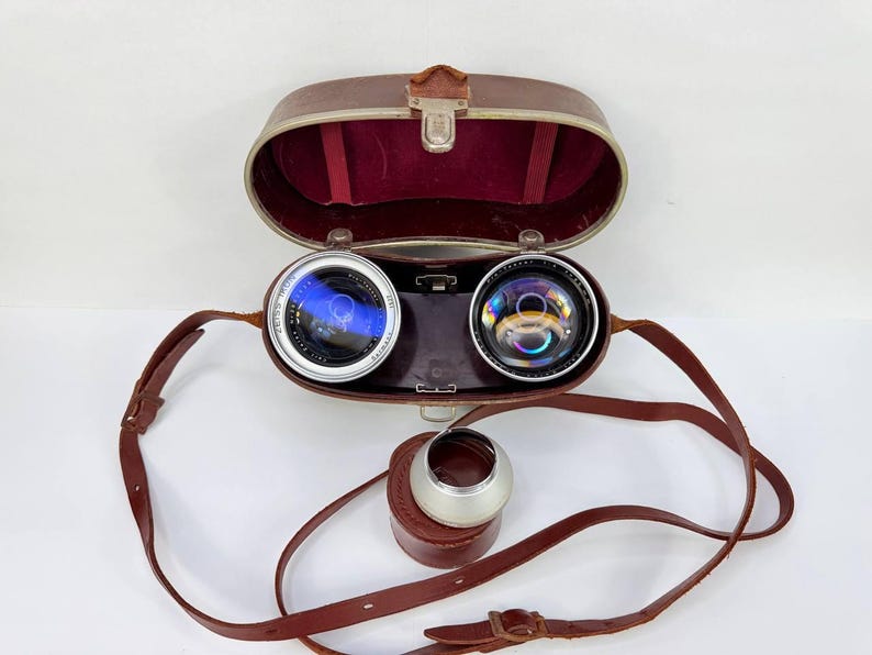 Puede incluir: Lentes de c&aacute;mara vintage con estuche y correa de cuero. Las lentes son plateadas y negras, con un interior azul reflectante. El estuche es marr&oacute;n con interior de terciopelo rojo. Tambi&eacute;n se incluye un parasol plateado y un peque&ntilde;o estuche de cuero.