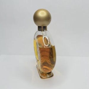 May include: A clear glass perfume bottle with a gold-colored spherical cap. The bottle is filled with a light amber liquid. The bottle has a unique, curved shape and sits on a white surface.