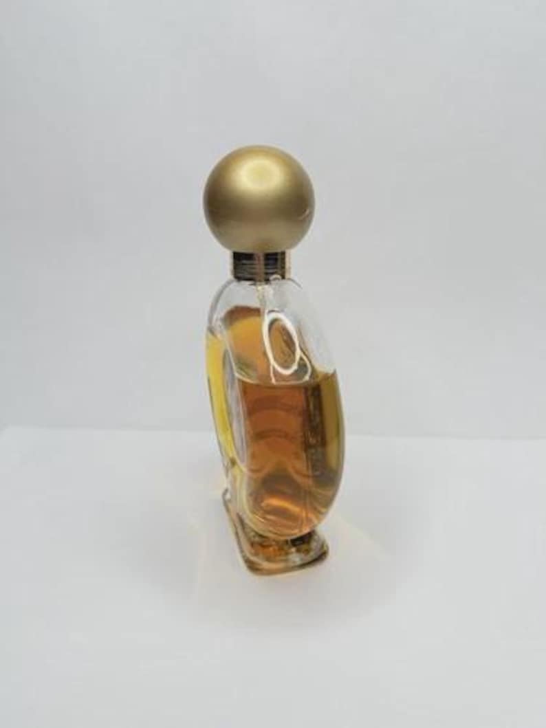 May include: A clear glass perfume bottle with a gold-colored spherical cap. The bottle is filled with a light amber liquid. The bottle has a unique, curved shape and sits on a white surface.