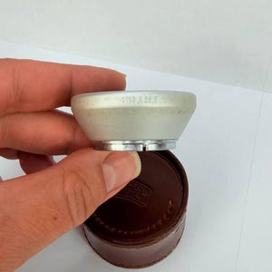 Puede incluir: Un componente de lente de c&aacute;mara plateado con el texto "1110 A 28,5" se sostiene sobre un estuche de cuero marr&oacute;n. El componente de la lente tiene forma c&oacute;nica con una base plateada. El estuche es redondo.