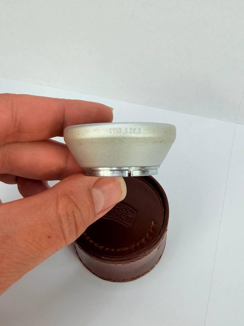 Puede incluir: Un componente de lente de c&aacute;mara plateado con el texto "1110 A 28,5" se sostiene sobre un estuche de cuero marr&oacute;n. El componente de la lente tiene forma c&oacute;nica con una base plateada. El estuche es redondo.