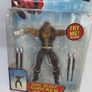 May include: A Dual Blast Shocker action figure from The Amazing Spider-Man, packaged in a clear plastic blister pack. The figure is brown and tan with silver accessories. The packaging includes the text "TRY ME! SEE BACK".