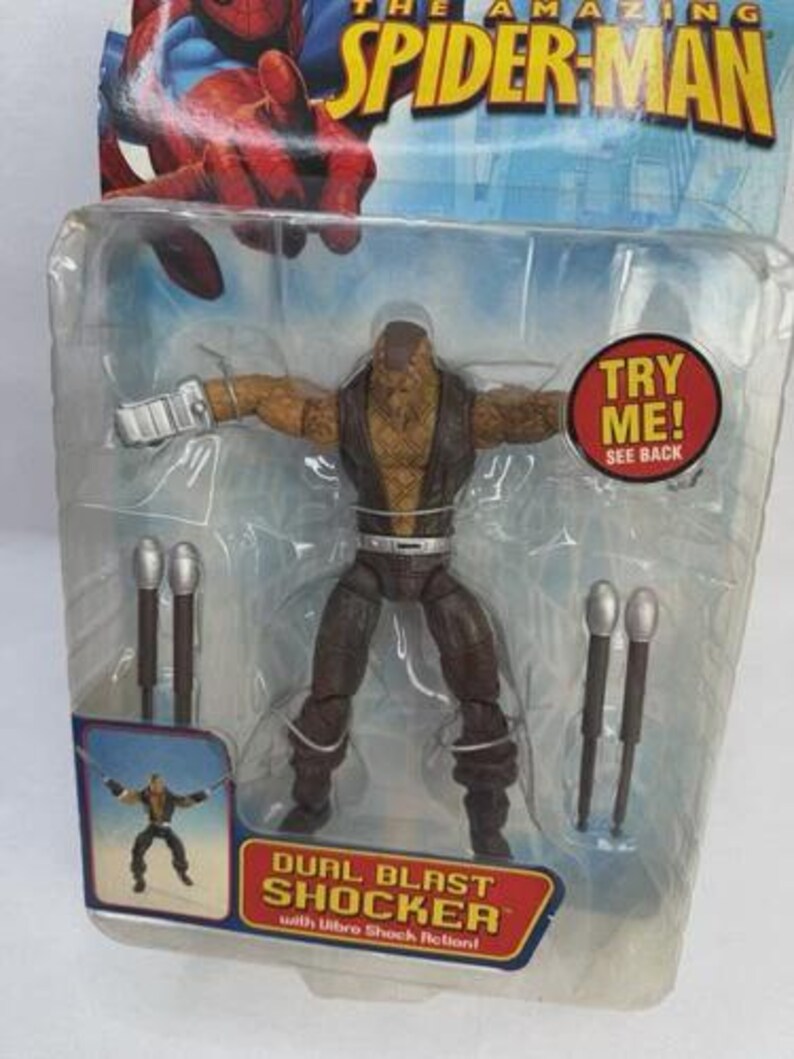 May include: A Dual Blast Shocker action figure from The Amazing Spider-Man, packaged in a clear plastic blister pack. The figure is brown and tan with silver accessories. The packaging includes the text "TRY ME! SEE BACK".