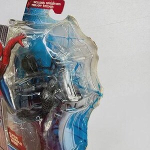 Peut inclure: Figurine Spider-Man dans un emballage plastique transparent. La figurine est noire et argent&eacute;e, avec une figurine Spider-Man rouge et bleue visible. L'emballage contient le texte "Includes Spider-Man Peel-Off Sticker."