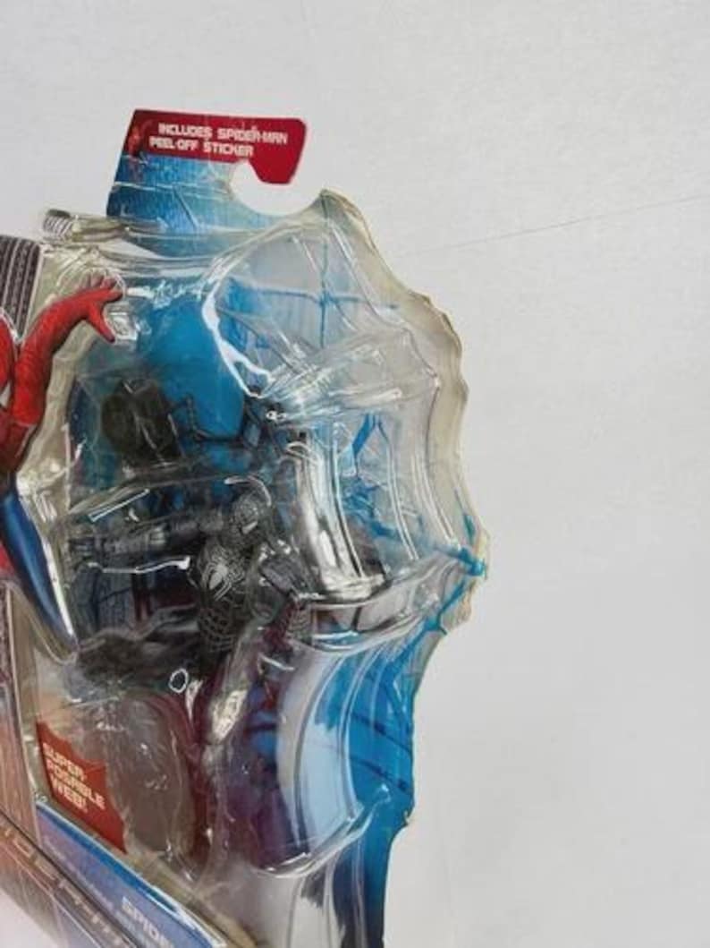Peut inclure: Figurine Spider-Man dans un emballage plastique transparent. La figurine est noire et argent&eacute;e, avec une figurine Spider-Man rouge et bleue visible. L'emballage contient le texte "Includes Spider-Man Peel-Off Sticker."