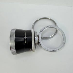 Puede incluir: Un objetivo de c&aacute;mara negro y plateado con el texto "Made in Germany" y "ZEISS IKON". Tambi&eacute;n hay dos anillos plateados presentes, uno con el texto "Germany" y el n&uacute;mero "1527".