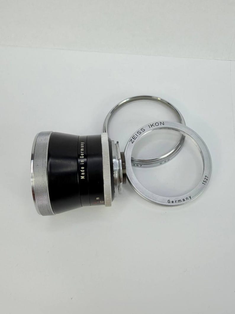 Puede incluir: Un objetivo de c&aacute;mara negro y plateado con el texto "Made in Germany" y "ZEISS IKON". Tambi&eacute;n hay dos anillos plateados presentes, uno con el texto "Germany" y el n&uacute;mero "1527".