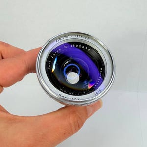 Puede incluir: Un objetivo de c&aacute;mara Zeiss Ikon plateado y negro con el texto "Zeiss Ikon" y "Germany" visibles. El objetivo tiene una superficie reflectante p&uacute;rpura y azul. El objetivo est&aacute; sostenido por una mano.