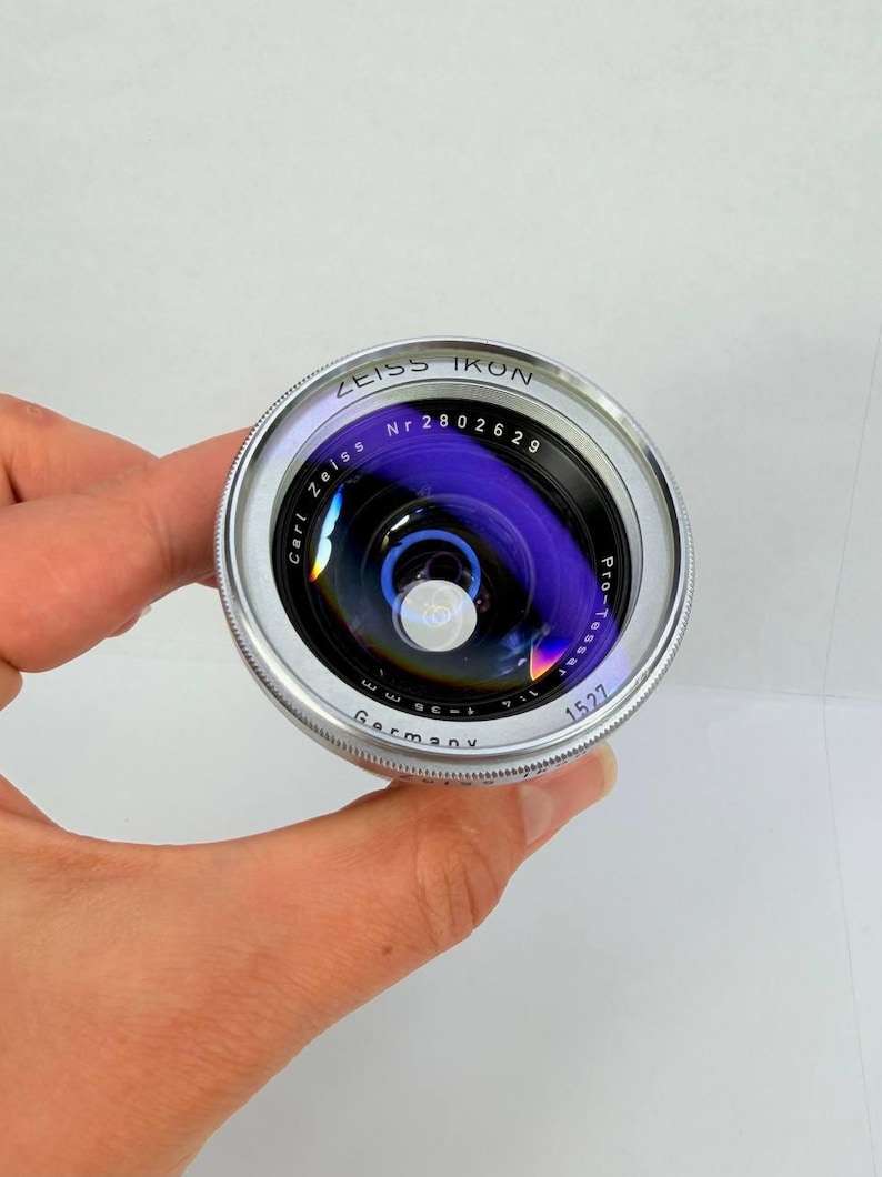 Puede incluir: Un objetivo de c&aacute;mara Zeiss Ikon plateado y negro con el texto "Zeiss Ikon" y "Germany" visibles. El objetivo tiene una superficie reflectante p&uacute;rpura y azul. El objetivo est&aacute; sostenido por una mano.