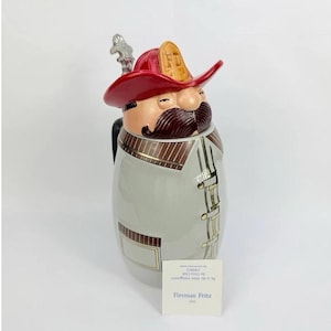 May include: A ceramic beer stein shaped like a firefighter with a red helmet, brown mustache, and a light gray uniform featuring gold trim. A small white card in front displays the text Fireman Fritz 1992, along with additional printed details.