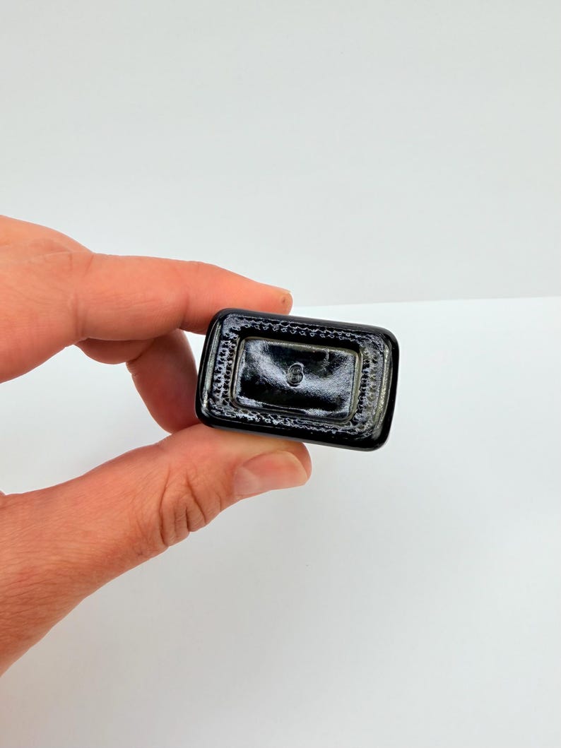 Pu&ograve; includere: Base rettangolare nera di bottiglia in vetro con superficie strutturata. La bottiglia &egrave; tenuta da una mano su sfondo bianco. La base presenta un piccolo marchio circolare.