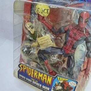 Peut inclure: Figurine de Spider-Man dans un emballage en plastique transparent. La figurine est rouge et noire, avec un motif de toile d'araign&eacute;e sur la poitrine. L'emballage comprend le texte "Spider-Man" et "Spider-Sense Spider-Man".