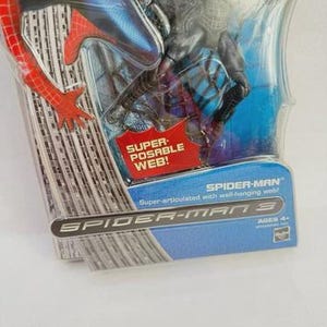 Peut inclure: Une figurine de Spider-Man dans un emballage en plastique transparent. La figurine est rouge et bleue, avec les mots "SUPER-POSABLE WEB!" et "SPIDER-MAN 3" visibles. L'emballage indique &eacute;galement "Super-articul&eacute; avec toile murale !" et "AGES 4+".