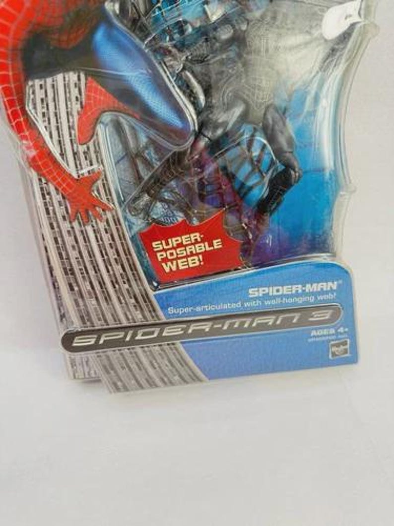 Peut inclure: Une figurine de Spider-Man dans un emballage en plastique transparent. La figurine est rouge et bleue, avec les mots "SUPER-POSABLE WEB!" et "SPIDER-MAN 3" visibles. L'emballage indique &eacute;galement "Super-articul&eacute; avec toile murale !" et "AGES 4+".