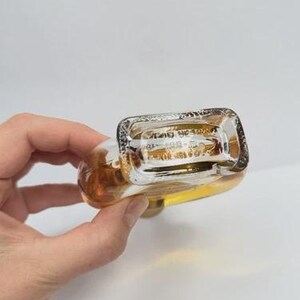 May include: A clear glass perfume bottle with a rectangular top and rounded body, filled with a light amber liquid. The bottle is held in a hand against a white background. The bottom of the bottle has etched text.
