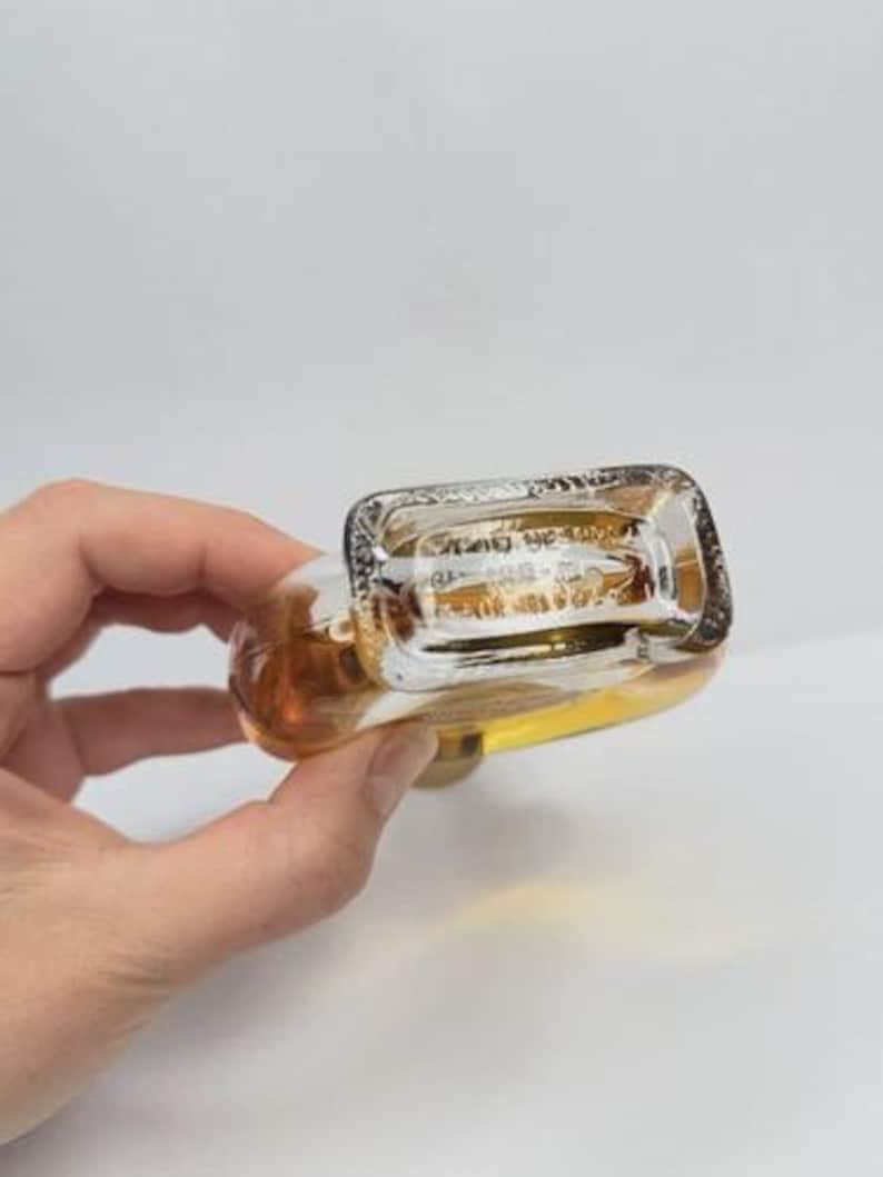 May include: A clear glass perfume bottle with a rectangular top and rounded body, filled with a light amber liquid. The bottle is held in a hand against a white background. The bottom of the bottle has etched text.