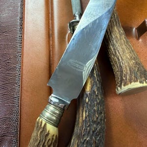 May include: A stainless steel knife with a bone handle and a silver bolster, resting on a brown leather surface. The knife has text on the blade. A sharpening steel and a second antler handle are also visible.