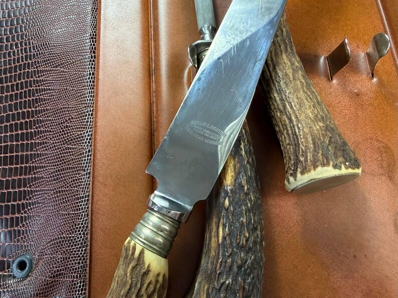 May include: A stainless steel knife with a bone handle and a silver bolster, resting on a brown leather surface. The knife has text on the blade. A sharpening steel and a second antler handle are also visible.