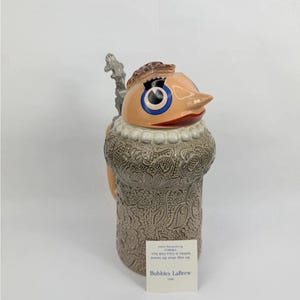 May include: A ceramic beer stein featuring a sculpted fish head lid with large blue eyes and a pointed snout. The body has a textured, tan, paisley-like pattern and a metal thumb-lift hinge. A white tag reads Bubbles LaBrew and includes text.