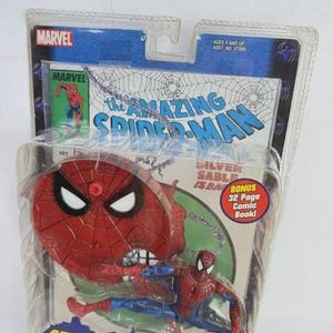 May include: A Spider-Man action figure from the Spider-Man Classics series, packaged in a clear plastic blister pack. The figure is red and blue, with a red spider-web-themed base. The packaging includes the Marvel logo and the text "The Amazing Spider-Man."