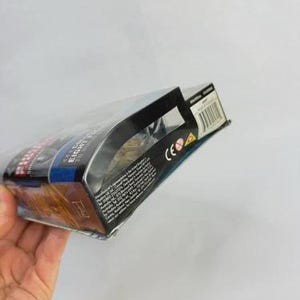 May include: A packaged action figure, held by a hand, with a clear plastic front. The box has a black top and sides with text and a barcode. The packaging features blue, red, and gold colors.