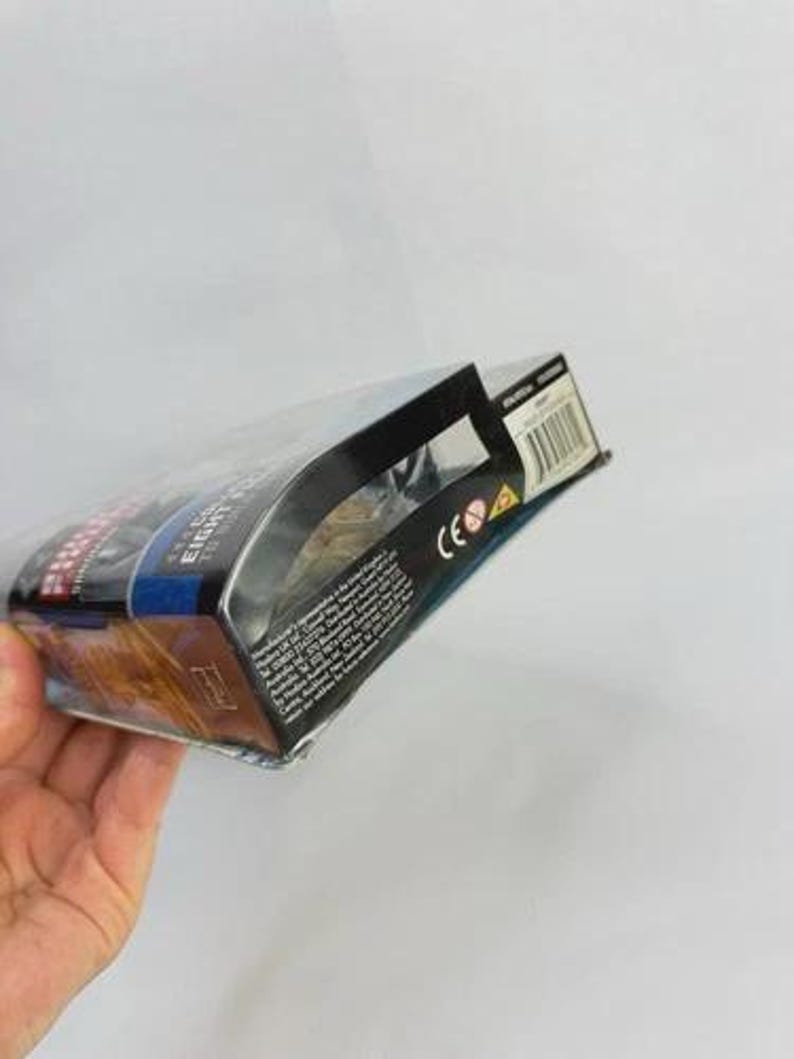 May include: A packaged action figure, held by a hand, with a clear plastic front. The box has a black top and sides with text and a barcode. The packaging features blue, red, and gold colors.