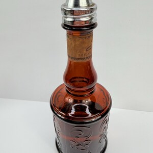 Puede incluir: Una botella vintage de vidrio &aacute;mbar con un tap&oacute;n de metal plateado. La botella tiene un patr&oacute;n floral decorativo y una etiqueta de corcho. La botella mide aproximadamente 15 cm de altura.