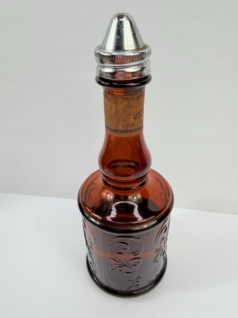 Puede incluir: Una botella vintage de vidrio &aacute;mbar con un tap&oacute;n de metal plateado. La botella tiene un patr&oacute;n floral decorativo y una etiqueta de corcho. La botella mide aproximadamente 15 cm de altura.