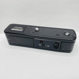 Puede incluir: Un accesorio de cámara negro Minolta Auto Winder G. El dispositivo tiene una parte superior texturizada, un interruptor de encendido y una pequeña luz roja. El texto "minolta AUTO WINDER G JAPAN" está impreso en la parte superior del dispositivo.