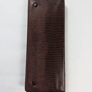 May include: A rectangular, dark brown leather wallet with a textured, reptile-like pattern. The wallet is closed and secured with two dark buttons. The design is simple and elegant.