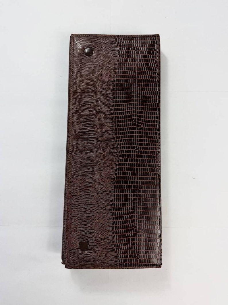 May include: A rectangular, dark brown leather wallet with a textured, reptile-like pattern. The wallet is closed and secured with two dark buttons. The design is simple and elegant.