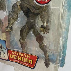 May include: Stealth Venom action figure in a clear plastic package. The figure is primarily olive green and brown, with detailed musculature. The packaging includes the text "TRY ME! SEE BACK" and "STEALTH VENOM with Symbiote Blast!"